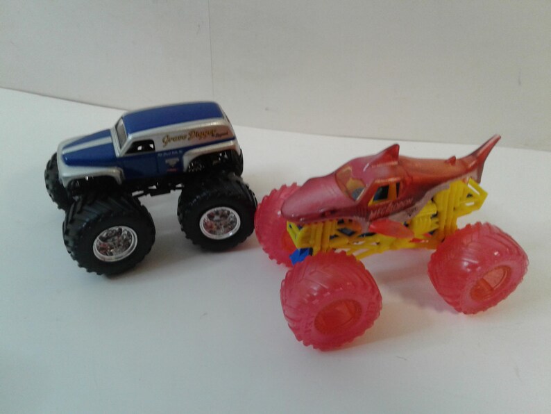 Może przedstawiać: Dwa zabawki monster truck. Jeden jest niebiesko-srebrny z czarnymi oponami. Drugi jest czerwono-ż&oacute;łty z czerwonymi kołami. Ciężar&oacute;wki zostały zaprojektowane z dużymi oponami i szczeg&oacute;łami.