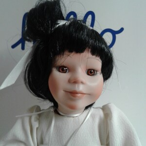 Pode incluir: Uma boneca de porcelana com cabelo preto preso em um coque, vestindo um vestido branco com uma fita branca ao redor do pesco&ccedil;o. A boneca tem olhos castanhos e um sorriso pintado.