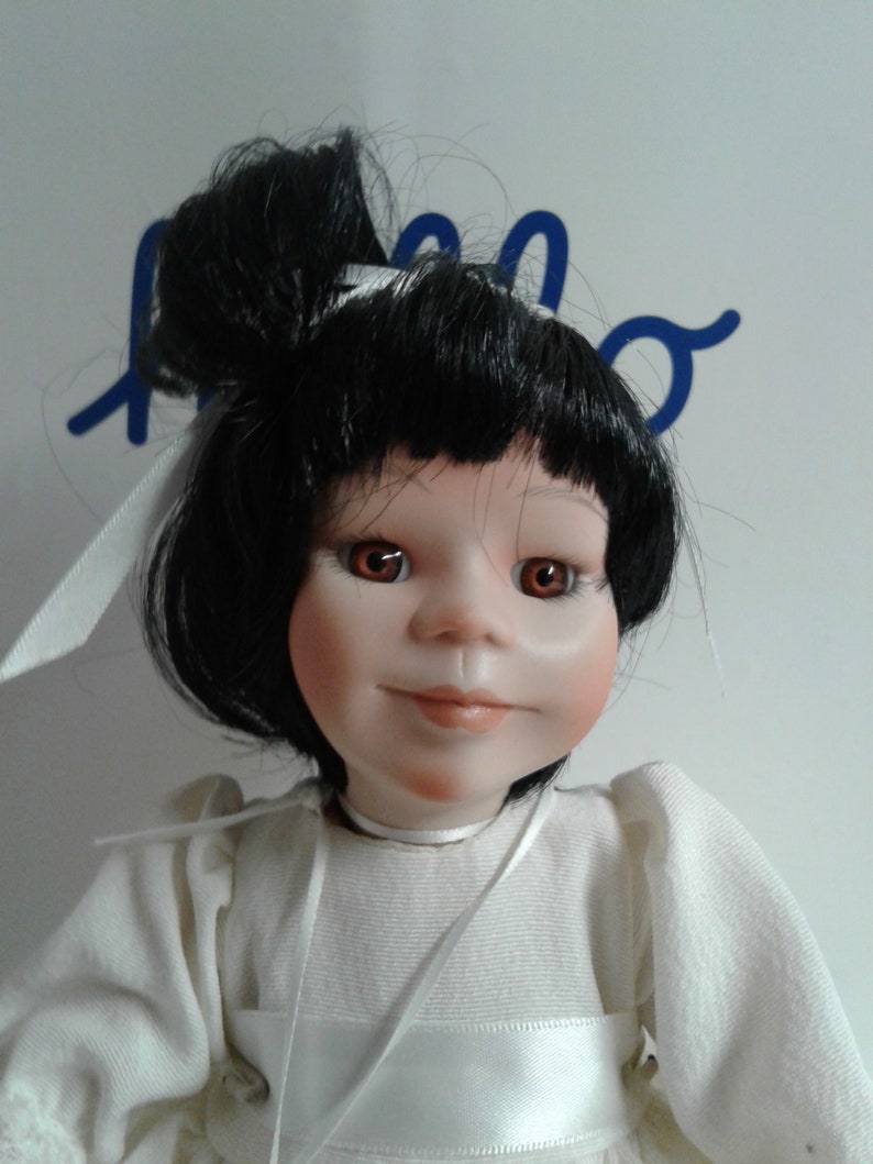 Pode incluir: Uma boneca de porcelana com cabelo preto preso em um coque, vestindo um vestido branco com uma fita branca ao redor do pesco&ccedil;o. A boneca tem olhos castanhos e um sorriso pintado.