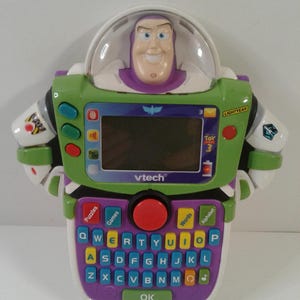 May include: A VTech Buzz Lightyear electronic learning toy featuring a QWERTY keyboard with colourful buttons, a central LCD screen, and a moulded plastic character head under a clear dome. The device includes labels for Puzzles, Games, Words, and Alphabet.