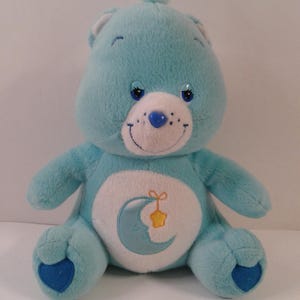 Könnte beinhalten: Ein hellblauer Plüschbär mit einem weißen Bauch, der ein Mond- und Stern-Design zeigt. Der Bär hat eine blaue Nase, Augen und Pfoten. Er hat einen Büschel blaues Fell auf dem Kopf und ein lächelndes Gesicht.