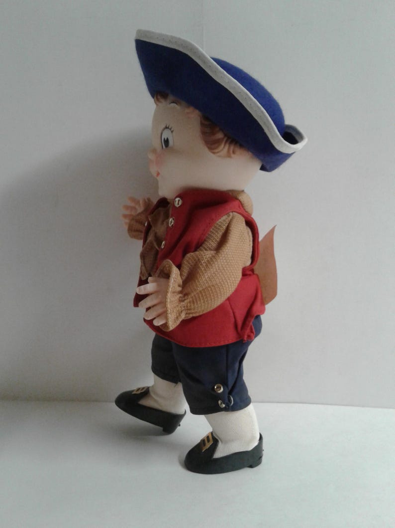 Pu&ograve; includere: Bambola vintage vestita con un abito in stile coloniale. La bambola indossa un cappello blu con finiture bianche, un gilet rosso e pantaloni blu scuro. Le scarpe della bambola sono nere con dettagli dorati. Il viso della bambola &egrave; dipinto.