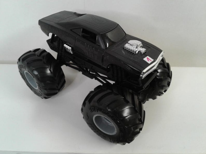 May include: Black monster truck toy with large tyres and a detailed engine. The toy car features a black body with silver engine detailing and a small red and white emblem on the front.