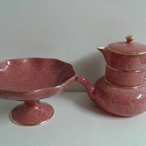 Puede incluir: Un juego de té rosa y dorado con un tazón a juego. La tetera está apilada con dos tazas y platillos. La tetera tiene un borde dorado y un diseño rosa moteado.