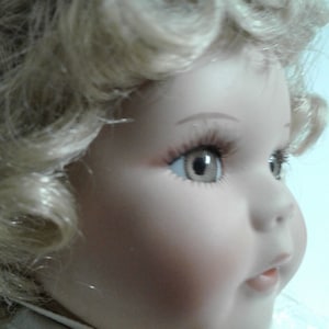 May include: A porcelain doll with blonde curly hair, blue eyes, and a pink and white dress. The doll has a sweet expression and is wearing a white bow.