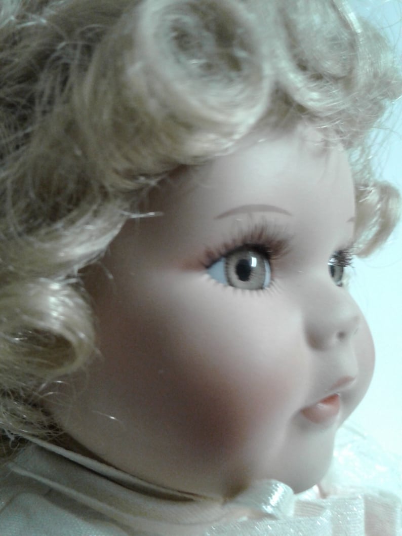 May include: A porcelain doll with blonde curly hair, blue eyes, and a pink and white dress. The doll has a sweet expression and is wearing a white bow.