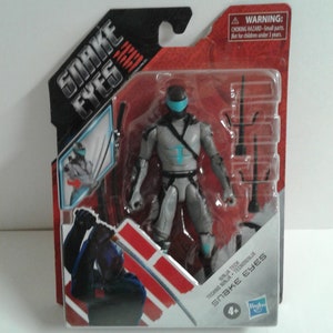 May include: A plastic package containing a gray and blue action figure of Snake Eyes from the G.I. Joe: Snake Eyes movie. The figure is holding a sword and has a blue visor. The package also includes a warning label for children under 3 years old. The package is labeled "Snake Eyes" and "Techno Ninja".