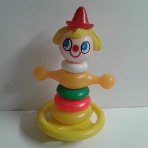 Puede incluir: Un juguete apilable de plástico vintage con forma de payaso. El payaso tiene un sombrero rojo, pelo amarillo y una cara sonriente. El juguete presenta anillos amarillos, rojos y verdes y mide aproximadamente 15 cm de alto.