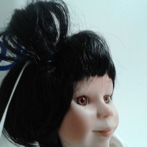 Pode incluir: Uma boneca de porcelana com cabelo preto preso em um coque, olhos castanhos e uma tez rosa claro. A boneca est&aacute; vestindo uma camisa branca.
