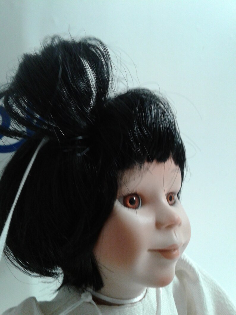 Pode incluir: Uma boneca de porcelana com cabelo preto preso em um coque, olhos castanhos e uma tez rosa claro. A boneca est&aacute; vestindo uma camisa branca.