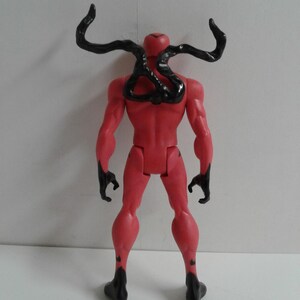 Pode incluir: Uma figura de a&ccedil;&atilde;o vermelha com tent&aacute;culos de simbionte pretos nas costas. A figura est&aacute; em pose com os bra&ccedil;os ao lado do corpo e as pernas ligeiramente afastadas.