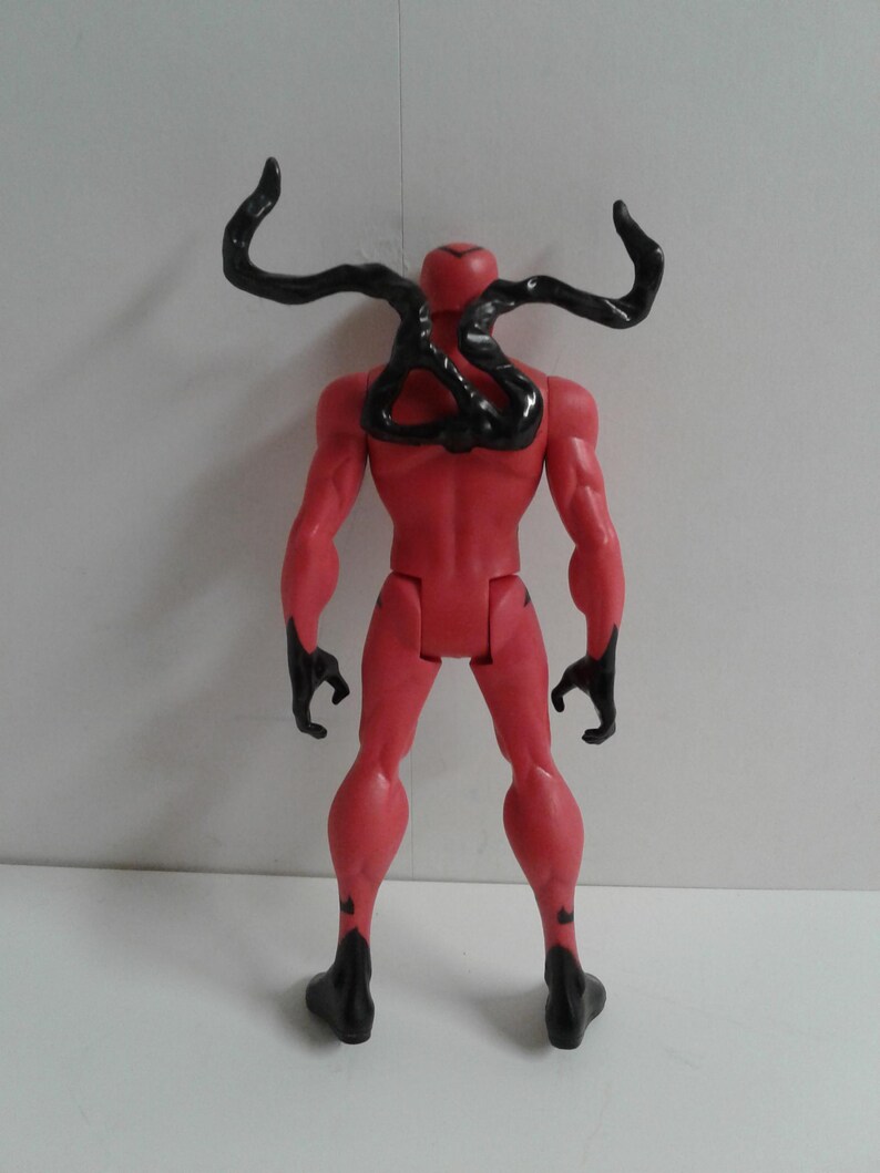 Pode incluir: Uma figura de a&ccedil;&atilde;o vermelha com tent&aacute;culos de simbionte pretos nas costas. A figura est&aacute; em pose com os bra&ccedil;os ao lado do corpo e as pernas ligeiramente afastadas.