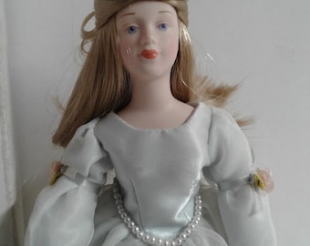 May include: A porcelain doll with blonde hair styled in a braid with flowers, wearing a light blue dress with a pearl necklace.