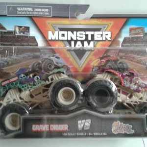 May include: A packaged Monster Jam toy set featuring two monster trucks: Grave Digger and El Toro Loco. The Grave Digger truck is green and black with a skull design, while El Toro Loco is red and purple. The package includes the text "Grave Digger VS El Toro Loco".