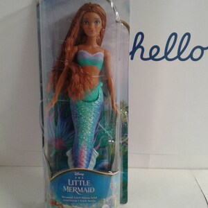 May include: A plastic package containing a Disney The Little Mermaid Ariel doll. The doll has long, red hair, a teal and purple mermaid tail, and a turquoise and purple top. The package has a blue background with images of Ariel and other sea creatures.