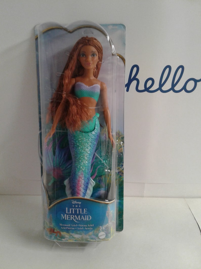May include: A plastic package containing a Disney The Little Mermaid Ariel doll. The doll has long, red hair, a teal and purple mermaid tail, and a turquoise and purple top. The package has a blue background with images of Ariel and other sea creatures.