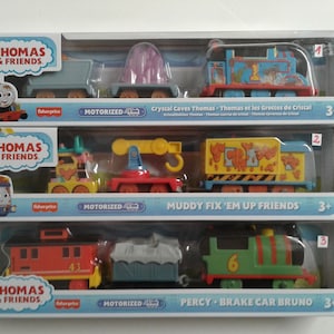 Könnte beinhalten: Drei Thomas & Friends Spielzeug-Züge, jeder mit einer motorisierten Lokomotive und einem Personenwagen. Das erste Set enthält eine blaue Lokomotive mit einem Kristallhöhlen-Design, das zweite Set enthält eine gelbe Lokomotive mit einem matschigen Reparatur-Design und das dritte Set enthält eine grüne Lokomotive mit einem Bremswagen und einem roten Personenwagen.