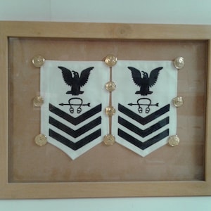Puede incluir: Insignias militares enmarcadas con dos parches blancos con galones negros y un emblema de águila. Botones dorados aseguran los parches dentro de un marco de madera, creando una exhibición de rango y servicio militar.