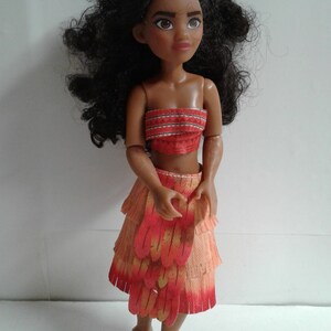 May include: A brown-skinned doll with long, curly black hair wearing a red and orange layered skirt with fringe and a red and white patterned top.