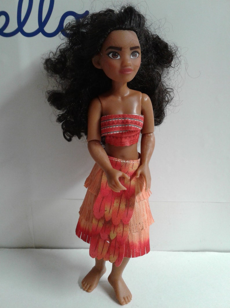 May include: A brown-skinned doll with long, curly black hair wearing a red and orange layered skirt with fringe and a red and white patterned top.