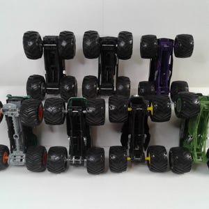 May include: A selection of toy monster trucks in various colours, including black, green, and purple. Each truck has large, black, knobbly tyres and a detailed chassis. The trucks are arranged in rows, showcasing their design.