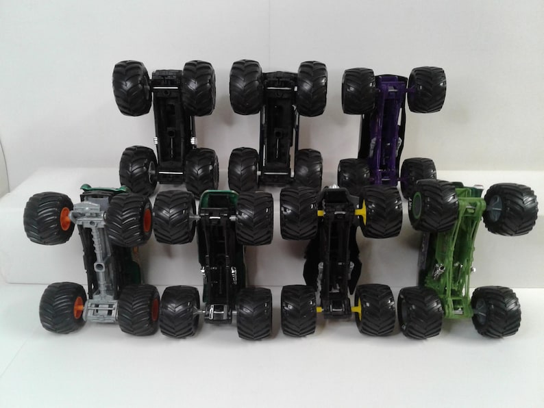 May include: A selection of toy monster trucks in various colours, including black, green, and purple. Each truck has large, black, knobbly tyres and a detailed chassis. The trucks are arranged in rows, showcasing their design.