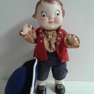 Pu&ograve; includere: Bambola vintage con espressione allegra, indossa un gilet rosso, una camicia marrone a balze e pantaloncini blu scuro. La bambola ha i capelli castani e le mani alzate. Un cappello blu con finiture bianche &egrave; appoggiato vicino.
