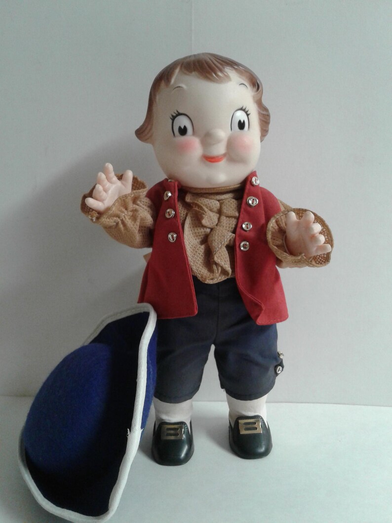 Pu&ograve; includere: Bambola vintage con espressione allegra, indossa un gilet rosso, una camicia marrone a balze e pantaloncini blu scuro. La bambola ha i capelli castani e le mani alzate. Un cappello blu con finiture bianche &egrave; appoggiato vicino.