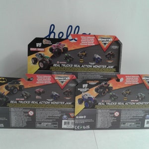 May include: Three Monster Jam toy packages are shown, each with different monster truck designs. The packaging includes the text "REAL TRUCKS! REAL ACTION! MONSTER JAM!" and the "VS" logo. The packages are mainly black, orange, and grey.