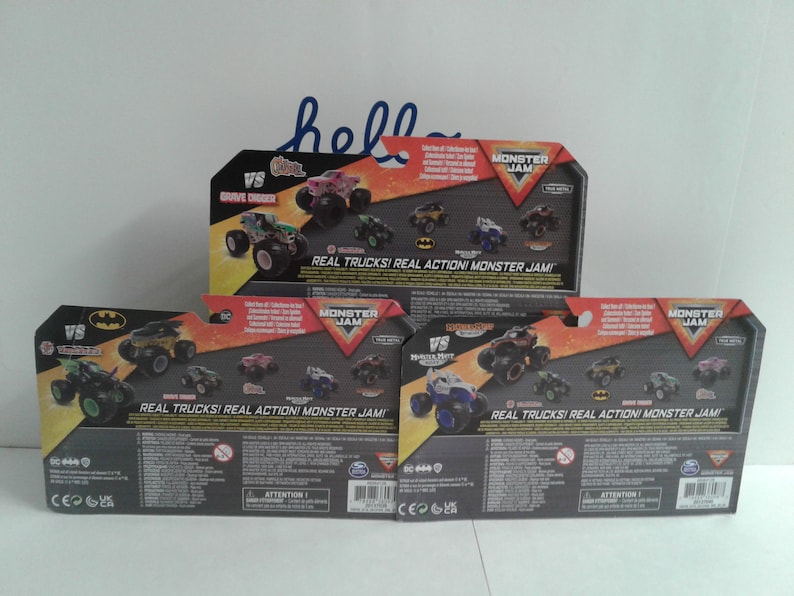 May include: Three Monster Jam toy packages are shown, each with different monster truck designs. The packaging includes the text "REAL TRUCKS! REAL ACTION! MONSTER JAM!" and the "VS" logo. The packages are mainly black, orange, and grey.