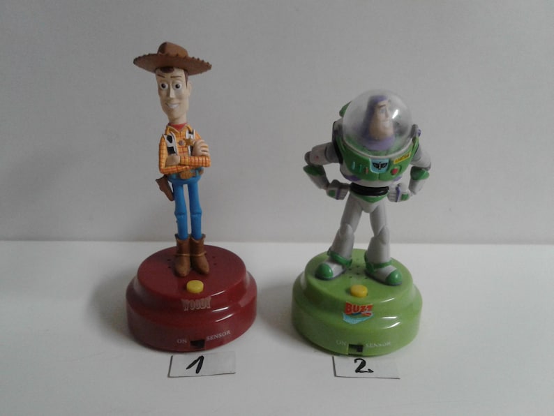 May include: Two character figurines from the animated film Toy Story. Woody, a cowboy, stands on a red base. Buzz Lightyear, an astronaut, stands on a green base. Both bases have a button and the text "ON SENSOR".
