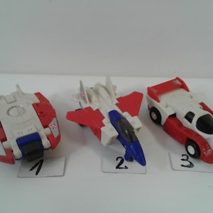May include: Three plastic toy vehicles in red, white, and blue. The first is a circular craft, the second is a fighter jet with wings, and the third is a race car. Each toy is labeled with a small white card numbered one, two, or three.