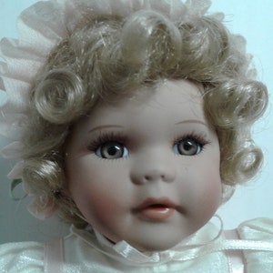 May include: A porcelain doll with blonde curly hair wearing a white dress with pink accents and a pink lace bonnet.