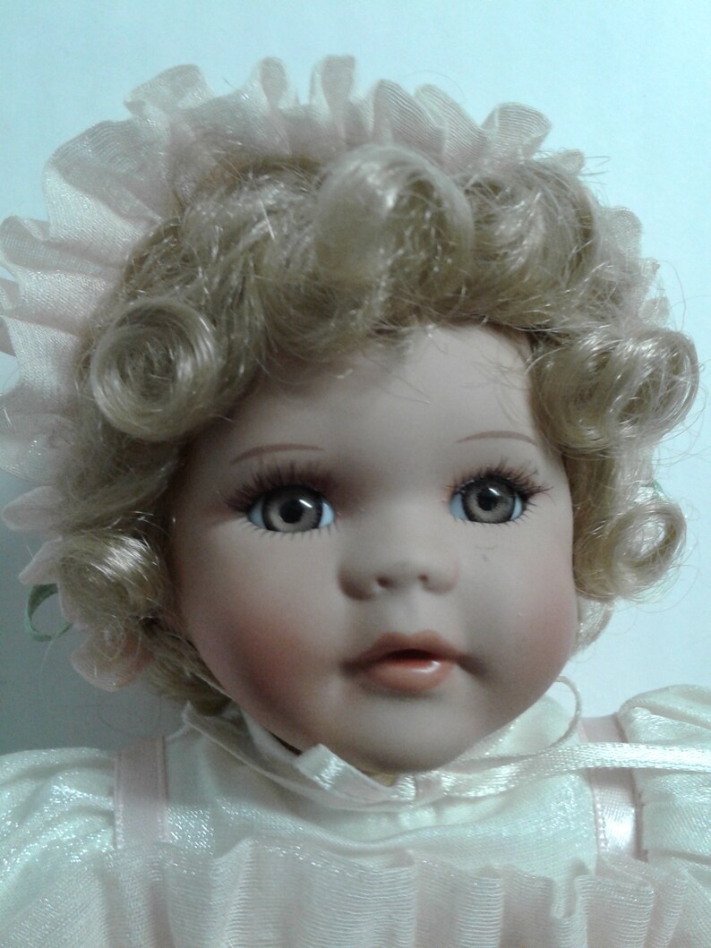 May include: A porcelain doll with blonde curly hair wearing a white dress with pink accents and a pink lace bonnet.