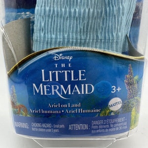 May include: A blue plastic doll of Ariel from Disney's The Little Mermaid, packaged in a clear plastic clamshell. The doll is wearing a blue dress and has long red hair. The packaging features the Disney logo and the words "The Little Mermaid" and "Ariel on Land". The packaging also includes a warning about choking hazards for children under 3 years old.