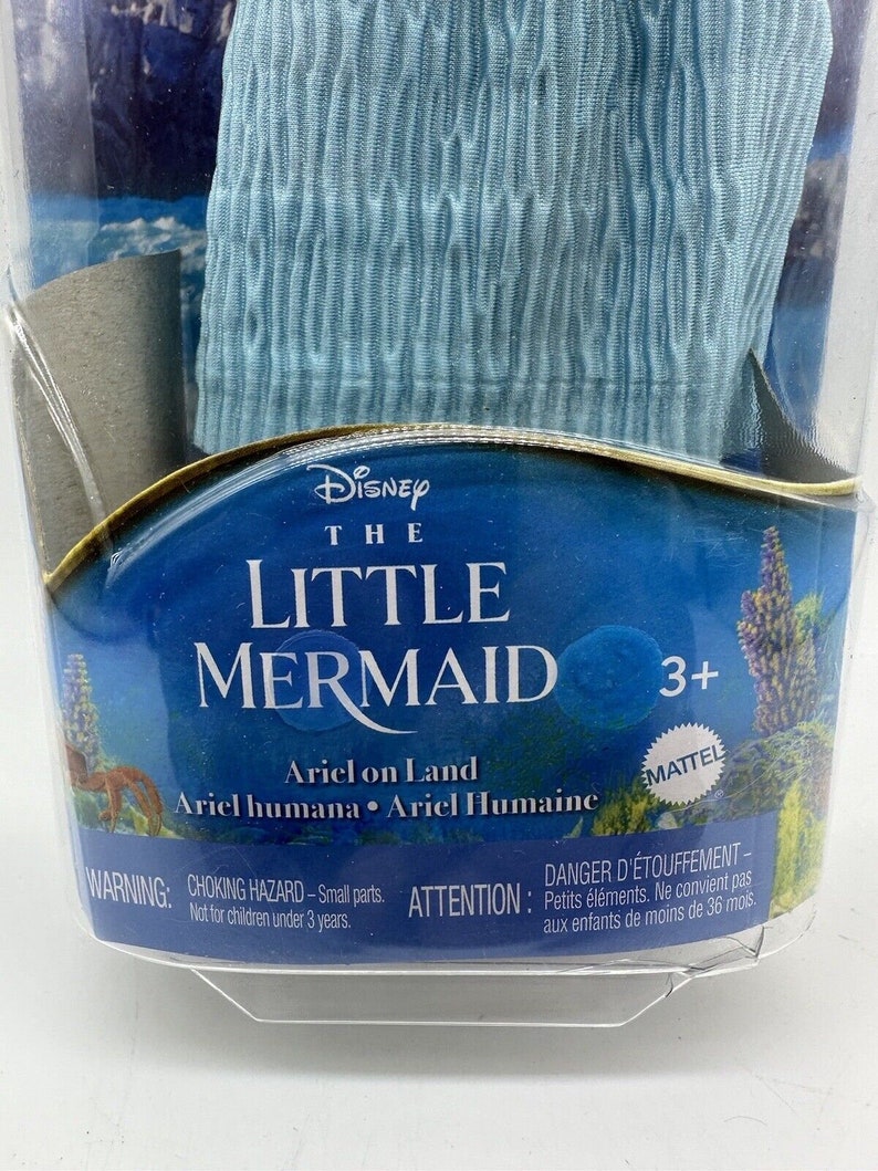 May include: A blue plastic doll of Ariel from Disney's The Little Mermaid, packaged in a clear plastic clamshell. The doll is wearing a blue dress and has long red hair. The packaging features the Disney logo and the words "The Little Mermaid" and "Ariel on Land". The packaging also includes a warning about choking hazards for children under 3 years old.
