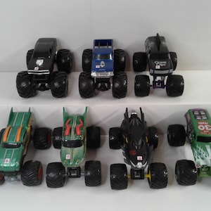 May include: A collection of seven toy monster trucks in various colours, including black, blue, green, and black. Each truck has large, oversized tyres and detailed designs on the body. The trucks are arranged on a white surface.