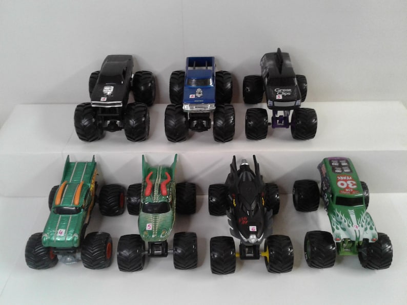 May include: A collection of seven toy monster trucks in various colours, including black, blue, green, and black. Each truck has large, oversized tyres and detailed designs on the body. The trucks are arranged on a white surface.