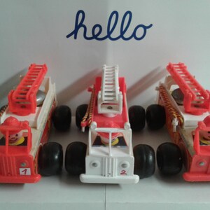 Könnte beinhalten: Drei alte Spielzeugfeuerwehrautos in Rot, Weiß und Beige. Jedes Auto hat eine rote Leiter, schwarze Räder und eine Feuerwehrfigur. Der Text "hello" steht in blauer Schreibschrift im Hintergrund. Die Nummern 1, 2 und 3 sind vorne.