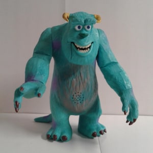 Puede incluir: Un juguete de peluche azul y morado del personaje Sulley de la película Monsters Inc. El juguete tiene una cabeza grande con dos cuernos, ojos grandes y una boca ancha. El juguete está de pie sobre una superficie blanca.