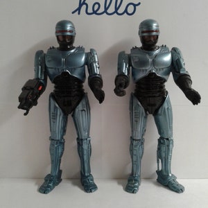 May include: Two metallic blue action figures of a robotic character, each with a black helmet and body armor. The figures are standing upright, with one holding a weapon. The word "hello" is written in cursive above them.