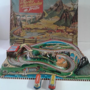 May include: Vintage "Rocky Mountains Train" toy set with a colorful landscape backdrop. The set includes a track with bridges, tunnels, and two small train cars in red and yellow. The box features a scenic illustration of mountains and a train.