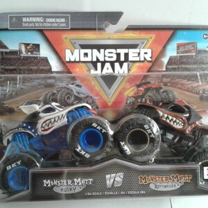 May include: A packaged Monster Jam toy set featuring two monster trucks: Monster Mutt Husky and Monster Mutt Rottweiler. The trucks are blue and black with detailed graphics and large tyres. The packaging includes the Monster Jam logo and a warning about small parts.