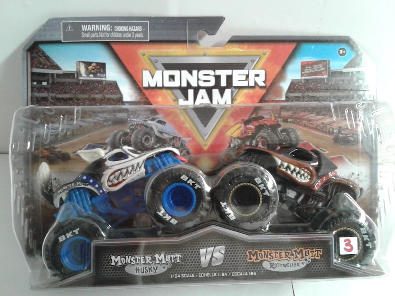 May include: A packaged Monster Jam toy set featuring two monster trucks: Monster Mutt Husky and Monster Mutt Rottweiler. The trucks are blue and black with detailed graphics and large tyres. The packaging includes the Monster Jam logo and a warning about small parts.