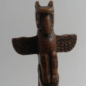 May include: A brown, carved wooden totem pole. The design features an owl-like figure at the top with outstretched wings. The totem pole has detailed carvings and a smooth finish, showcasing traditional Native American art.
