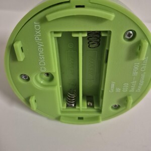 May include: A light green plastic device with a battery compartment. The compartment is designed for two AAA batteries. The device has the Disney/Pixar logo and text that reads "Gemmy HF 0310 Batch-HF001 Dongguan, China."