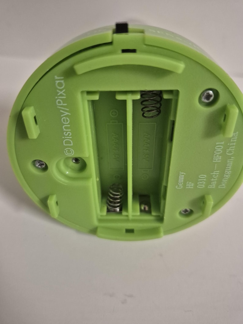 May include: A light green plastic device with a battery compartment. The compartment is designed for two AAA batteries. The device has the Disney/Pixar logo and text that reads "Gemmy HF 0310 Batch-HF001 Dongguan, China."