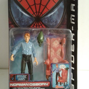 May include: A plastic package containing a Norman Osborn action figure from the Spider-Man movie. The figure is wearing a blue shirt, black pants, and a green goblin mask. The package also includes a red armchair and a small picture of the figure sitting in the chair. The text on the package reads "Norman Osborn" and "with Talking Goblin Mask (English)".