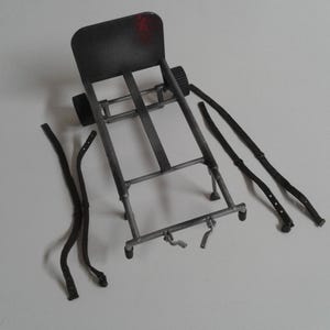 May include: A metal frame with a black back panel and four black straps. The frame has two wheels and two legs. The straps are used to secure items to the frame.