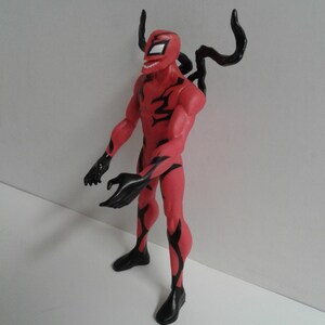 Pode incluir: Figura de a&ccedil;&atilde;o vermelha e preta de um super-her&oacute;i com um traje simbi&oacute;tico. A figura tem uma express&atilde;o amea&ccedil;adora e est&aacute; em pose com os bra&ccedil;os estendidos.
